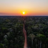 A photo taken from high above a tropical forest of green trees, stretching out to the horizon where the sun is just about to set