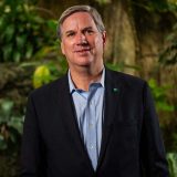 A person with short gray hair in a navy suit jacket and blue button-up shirt poses for a photo in a tropical environment