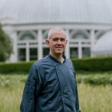 Head shot of Jeff Haskell wearing a blue chef shirt standing in front of tall pale yellow grass with a victorian appearing round glasshouse in the background.