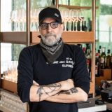 Head shot of Johnny Swet wearing a black shirt with a black bandana around his neck, and a black baseball hat. Grey short haired beard and standing in front of a brown open shelf with many wine glasses on each of the two levels.