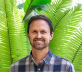 Head shot of Colin Khoury wearing a blue plaid shirt with brown short facial hair in a beard and goatee, standing in front of tall green palm frawns.