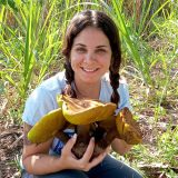 A person with brown pigtails kneels in the grass, holding a collection of enormous brown and yellow mushrooms