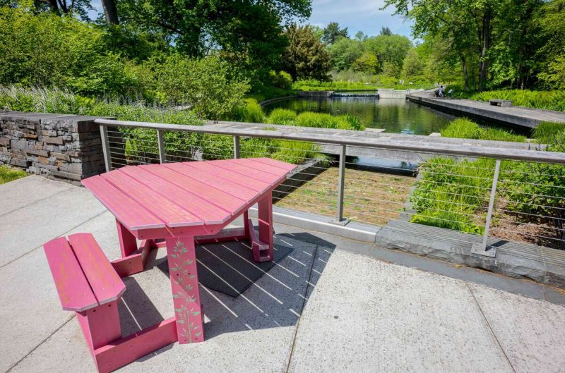 A red, triangular picnic table sits in the sun on a deck overlooking a lush garden space, with a water feature visible in the background