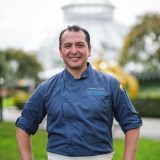 Head shot of Nelson Sivichay wearing a blue chef shirt with brown short cropped hair and a blurred background.