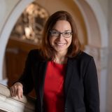 Headshot of Dr. Katherine Hayhoe wearing a black blazer, red shirt, with shoulder length brown hair, blue glasses and is standing with an arm bent on a marble staircase with a marble arch in th background.
