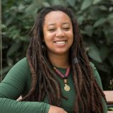 A person in a long-sleeved green shirt with long dreadlocks poses for a photo in an outdoor setting