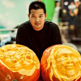 A person with short black hair and a black shirt poses for a photo between two carved pumpkins