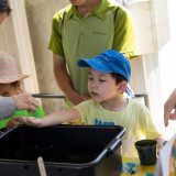 A child reaches for seeds over a bucket of soil