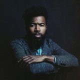 A person with short black hair and a dark beard poses for a photo, leaning over a chair while wearing a gray jacket