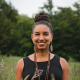 A photo of a woman in a black shirt and long beaded necklace, with her hair in a bun, posing for a photo outdoors