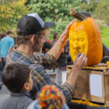 A person in a trucker hat carves a face into an orange pumpkin as a child watches