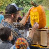 A person carves a face into an orange pumpkin as a child watches