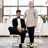 Two people in contrasting black, white, and beige outfits pose for a photo in a creative studio