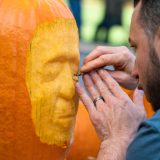 A person uses a small tool to carve the uncanny likeness of a human face into a large orange pumpkin