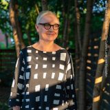 A person with short white hair and black glasses poses for a photo in a black and white striped dress