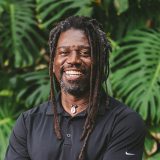 A person with long dark hair, a beard, and a black shirt poses for a photo in front of lush plants