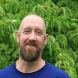 A person with a brown beard and blue shirt poses for a photo with plants in the background