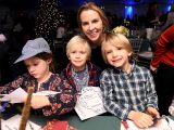 Four people looking at the camera smiling in holiday attire cutting out a craft with a blurred christmas tree in the background.