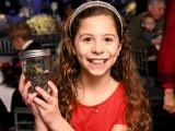 A young person wearing a red top and a sparkly head band with long curly brown hair and a toothy upturned mouth, holds a glass mason jar filled with soil and small plants.