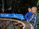 A person in a blue and white plaid shirt holds a small child wearing a blue shirt with blark writing to a brown train track with a miniture blue train that is driving by.