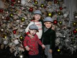 Three young children dressed in festive attaire of trains and green velvet jackets, with train conductor hats, stand in front of white frosted holiday trees with shiny green red and white decorative balls on them.