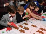 Four children dressed in festive attire wearing jackets and bow ties, and a dress, holding red, green, and blue cake decorations above brown gingerbread shapped cookies on a white table cloth.