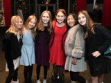 Six young children dressed in festive attire of dresses, and tights stand together in a posed photo with teethy upturned mouths.
