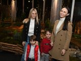 Two people in white tops, and jackets hold the hands of smaller children in red tops in front of model trains.