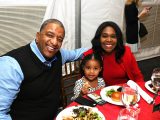 Two people, one wearing a black vest over a blue and white shirt, the other in a red top and sweater sit with a small child between them wearing a balck and white polka dot top. They have food on the table with a red cloth.