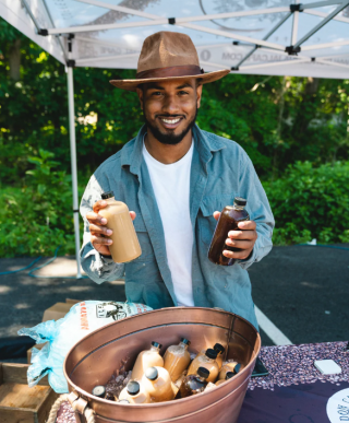 A person in a brimmed hat and denim shirt holds up bottles of iced coffee on a sunny day