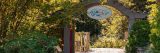 Wide view of an entry gate to the Everett Children's Adventure Garden, with a brown wooden archway, covered in green foliage up along the sides. Sun shines on the path below with an open view into more green space.