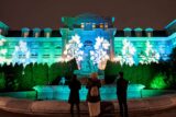 Three people in winter clothes look up at a classical building lit brightly with blue, green, and white lights in botanical patterns