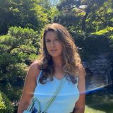 A person in a blue shirt with long, dark hair poses for a photo in a garden setting on a sunny day