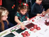 Three small children in festive attire wearing dresses and bows are holding red potatos cutin half and using one side to press onto a green square.