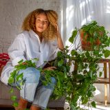 A person with curly blonde hair and a white shirt poses with a vining green plant in the sunlight