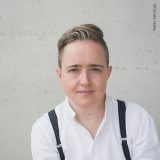 A person with short gray-brown hair, a white shirt, and black suspenders poses for a photo