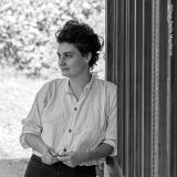 A person in a loose white shirt and black pants leans against a window while posing for a black and white photo