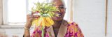 A person with short blonde hair and a bright pink blazer holds up a crafted yellow and green flower in a brightly lit room