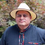 A person in a blue buttoned shirt and white brimmed hat poses for a photo outdoors