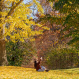 a person in a brown jacket and jeans sitting on the grass pointing up to the trees above with yellow and green leaves