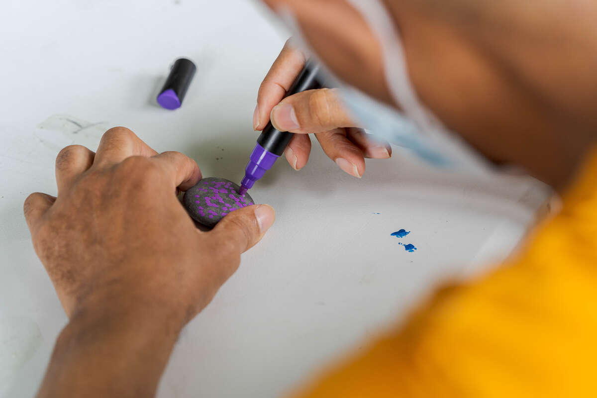 A person uses a purple paint pen to draw flowers on a gray rock
