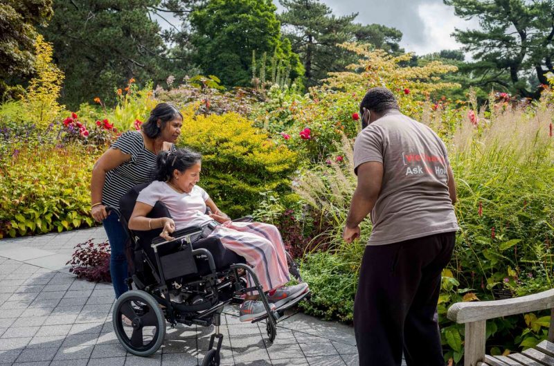 A person in a dark dress pushes another person in pink clothing, sitting in a wheelchair, as they explore a green garden space with a third person in a brown shirt and black pants
