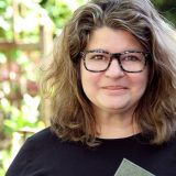 A person with wavy hair and glasses smiles softly at the camera, with an unfocused background of green plants.