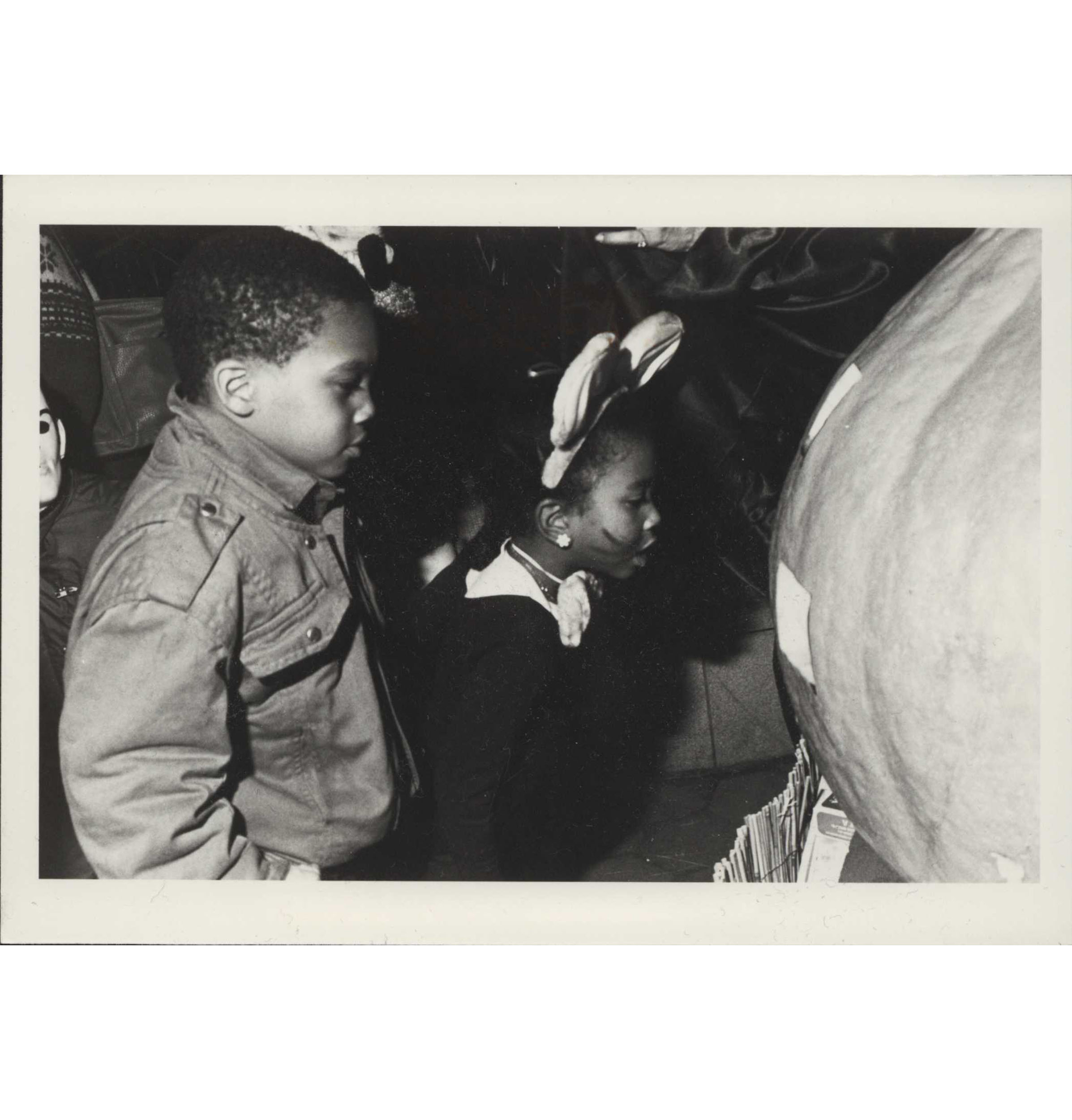 A black and white photograph of two children looking at a large pumpkin. One wears a costume with bunny ears and whiskers painted on her face.
