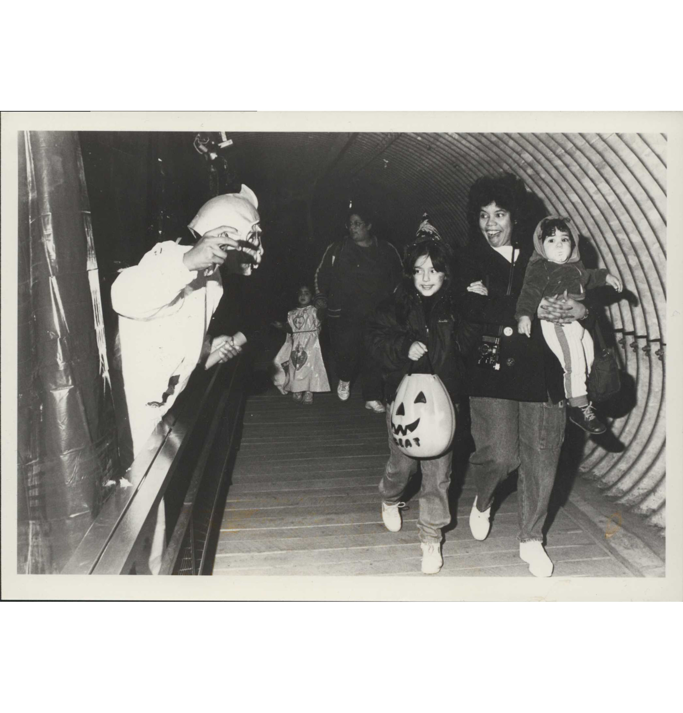 A black and white photograph of a smiling woman holding a small child and another child holding the woman's hand with a jack-o-lantern bag in her other. On the left, a person dressed in a costume jumps out from behind curtains to suprise passerby.