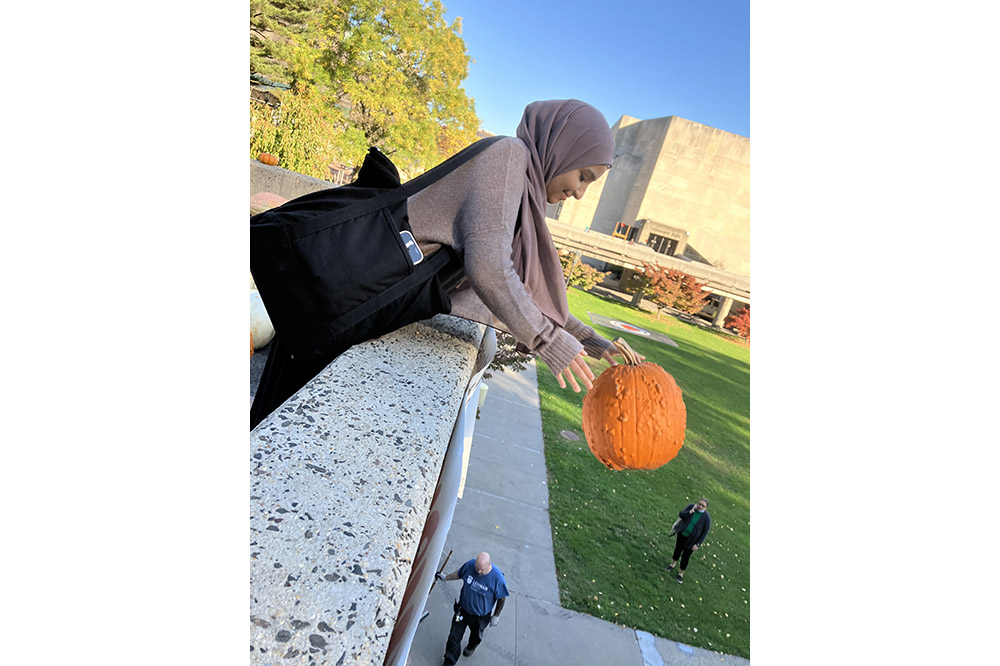A person in a beige shirt and hijab drops an orange pumpkin off a rooftop on a sunny day