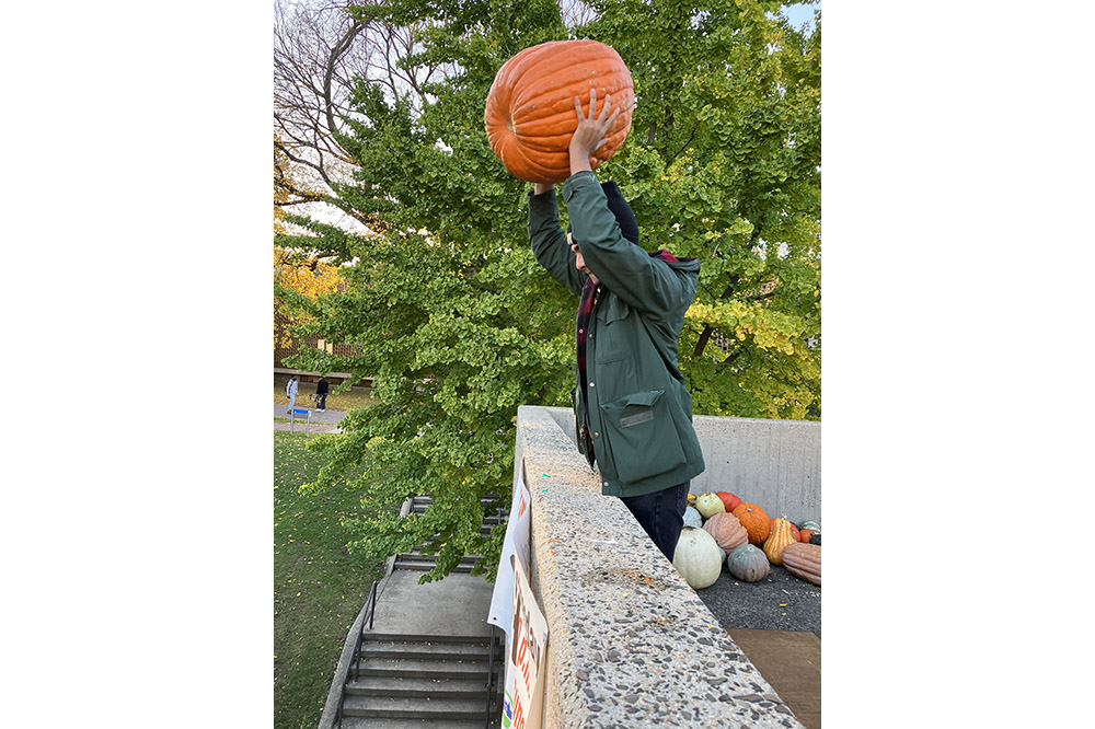 A person in a green sweatshirt lifts a big orange pumpkin over their head before launching it off a balcony