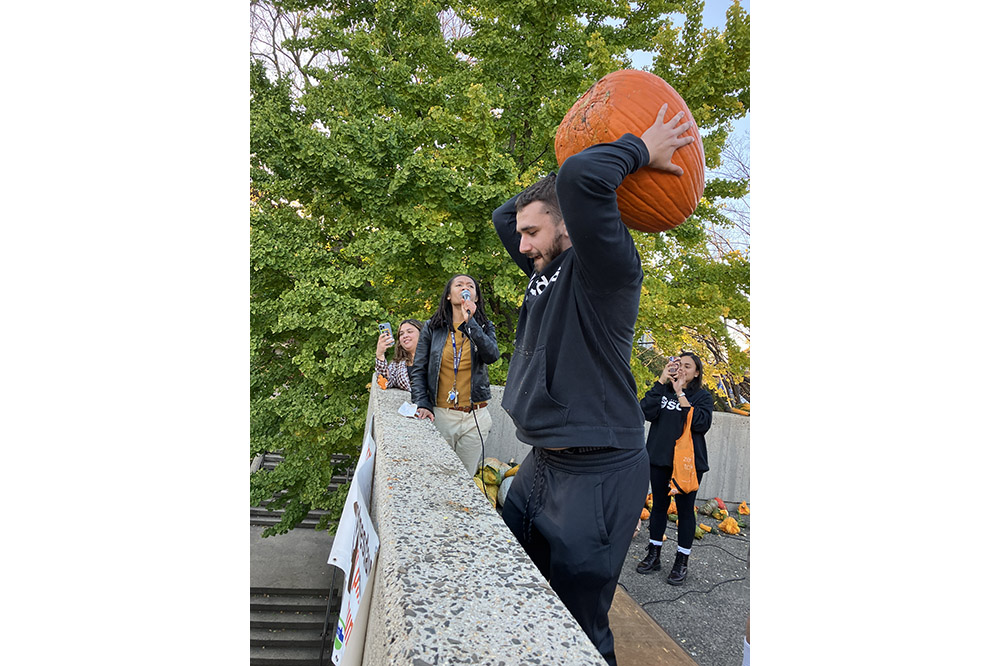 A person in a black sweatshirt winds up a throw with an enormous orange pumpkin