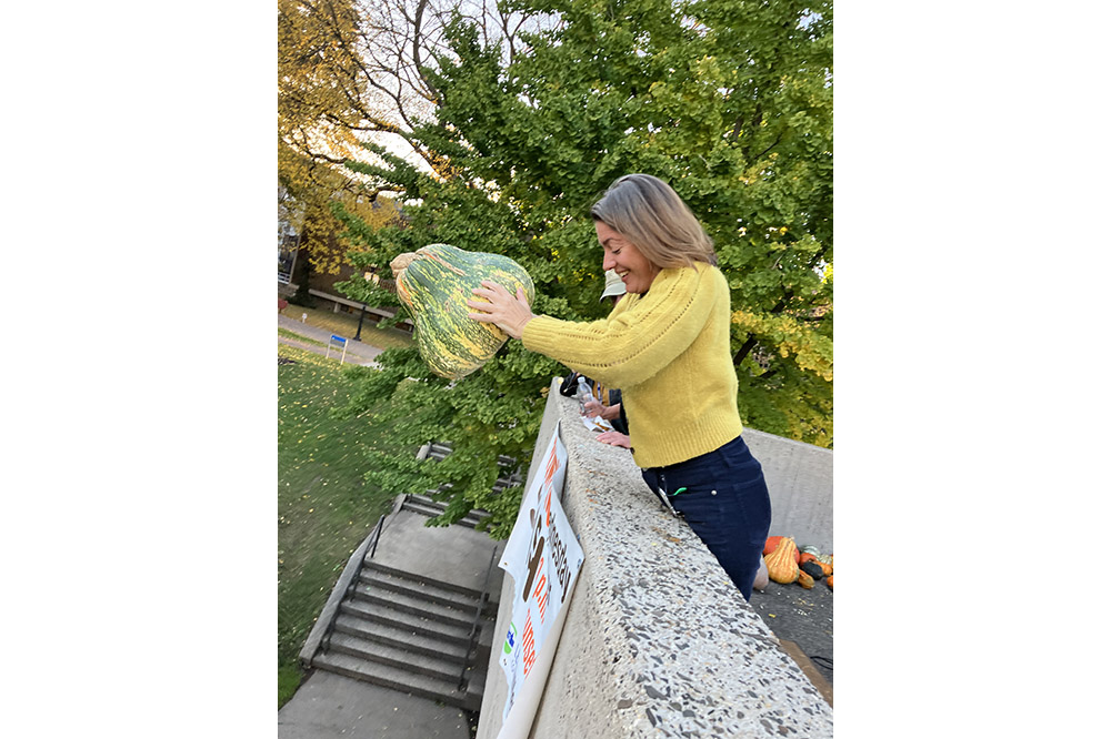 A person in a yellow sweater drops a green and white pumpkin off a roof