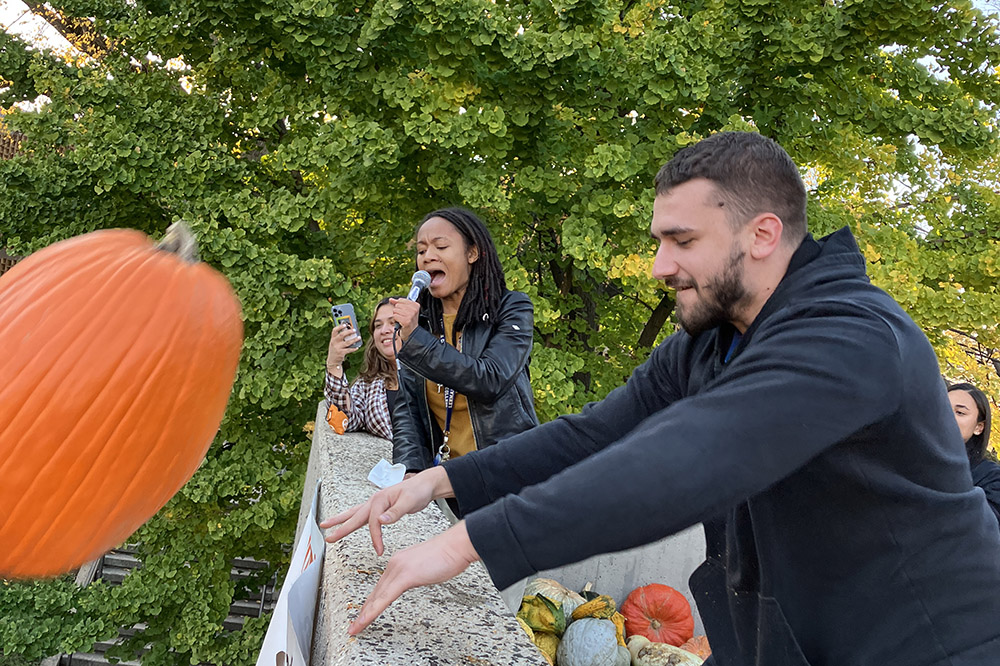 A person in a black sweatshirt throws a large orange pumpkin off a roof as an announcer with a microphone narrates in the background